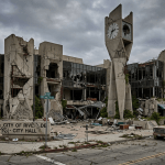 Damaged city hall building with large cracks and broken windows surrounded by debris and an old truck